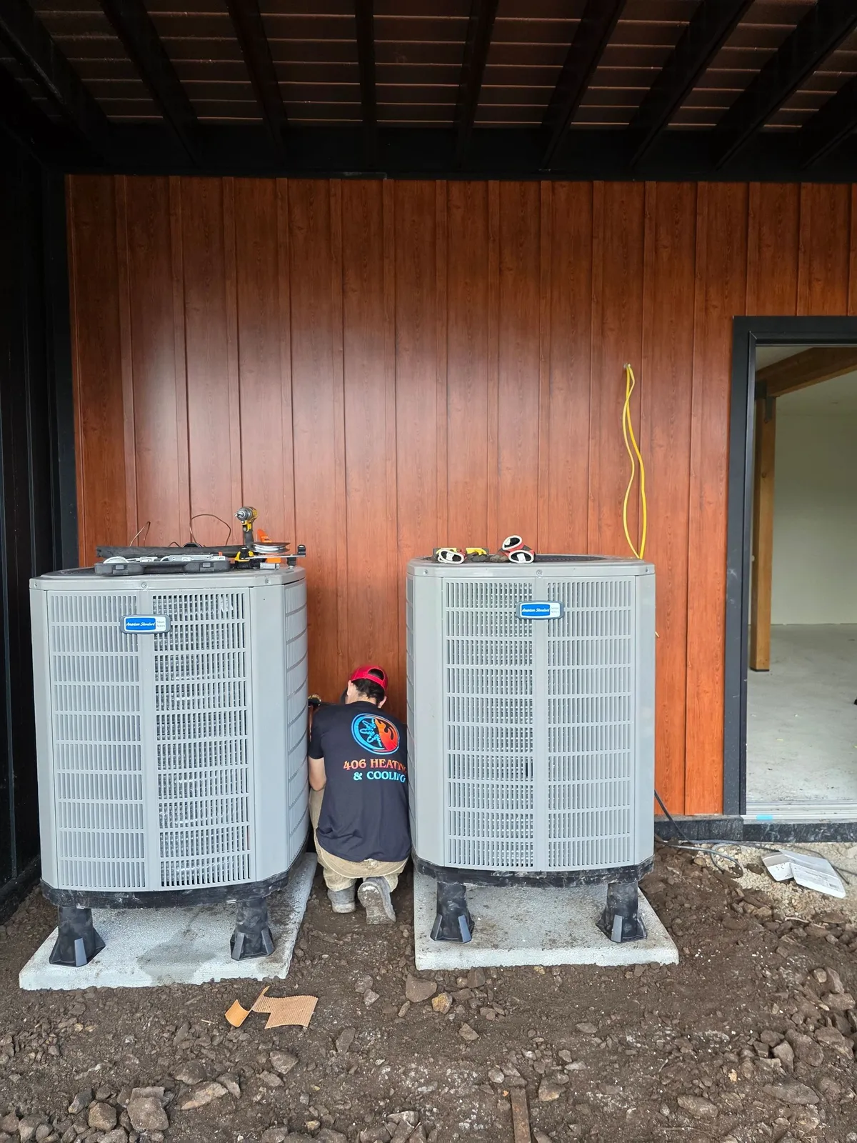Two American Standard condensers set on concrete pads on equipment feet at a new construction home with wood panel siding, and a 406 Heating and Cooling technician in a branded company shirt working between them